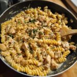 A close-up of creamy garlic parmesan chicken pasta with fusilli noodles and chopped chicken in a black pan, garnished with parsley.