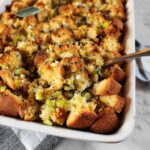 Close-up of a golden-brown Thanksgiving stuffing in a white baking dish, with visible chunks of bread, celery, and herbs.