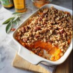 A close-up of a Thanksgiving side dish: sweet potato casserole with a pecan and oat crumble topping in a white baking dish.